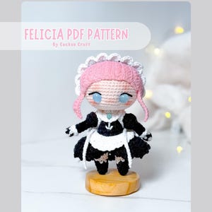 May include: A crocheted doll with pink hair, a white lace-trimmed headpiece, and a black and white maid outfit. The doll has light blue eyes and stands on a round wooden base. The text FELICIA PDF PATTERN By Cuckoo Craft is visible.