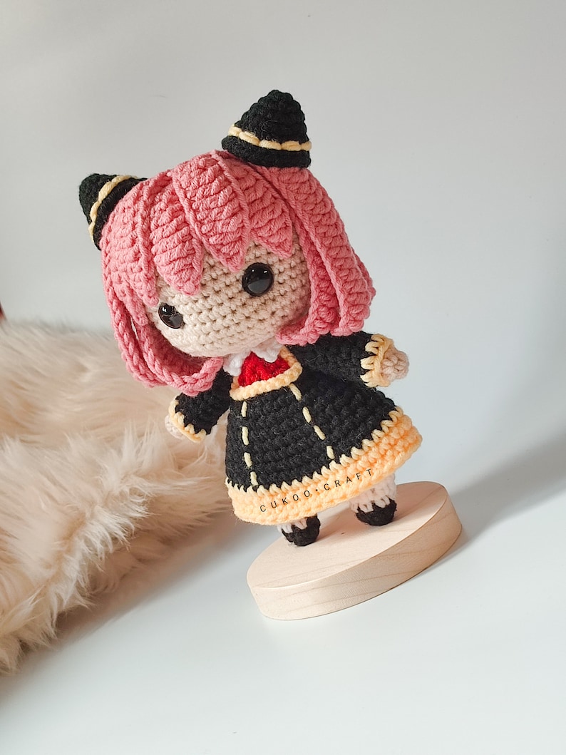 K&ouml;nnte beinhalten: Eine geh&auml;kelte Puppe mit rosa Haaren, schwarzen Ohren und einem schwarzen und gelben Kleid. Die Puppe steht auf einem Holzsockel. Die Puppe ist handgefertigt und hat ein s&uuml;&szlig;es und fantasievolles Design.