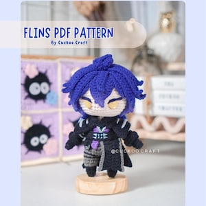 Könnte beinhalten: Handgefertigte Häkelpuppe mit blauen Haaren und einem schwarzen Outfit. Die Puppe steht auf einem Holzsockel. Der Text "FLINS PDF PATTERN" steht oben im Bild, darunter "By Cuckoo Craft". Die Puppe hat gelbe Augen.