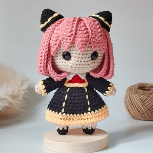 K&ouml;nnte beinhalten: Eine geh&auml;kelte Puppe mit rosa Haaren, schwarzen Ohren und einem schwarzen und gelben Kleid. Die Puppe steht auf einem Holzsockel. Die Puppe ist handgefertigt und hat einen niedlichen, verspielten Look.