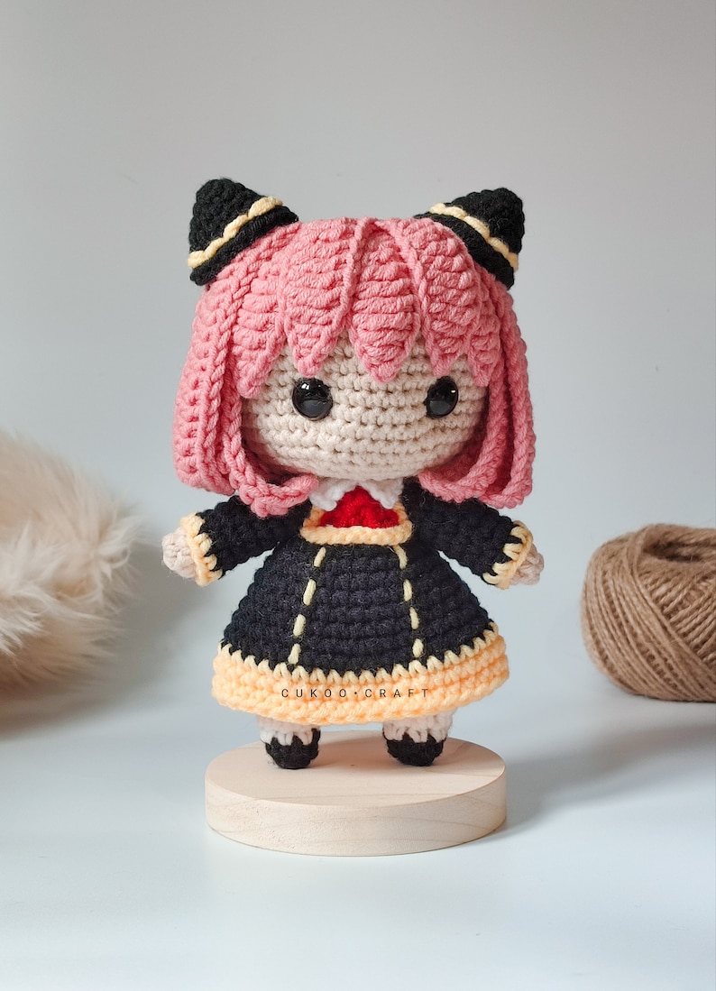K&ouml;nnte beinhalten: Eine geh&auml;kelte Puppe mit rosa Haaren, schwarzen Ohren und einem schwarzen und gelben Kleid. Die Puppe steht auf einem Holzsockel. Die Puppe ist handgefertigt und hat einen niedlichen, verspielten Look.