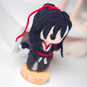 May include: Handmade crochet doll featuring black hair and a black outfit with red detailing. The doll stands on a small wooden base. It has large black eyes and a red string in its hair. The doll is a handcrafted item.