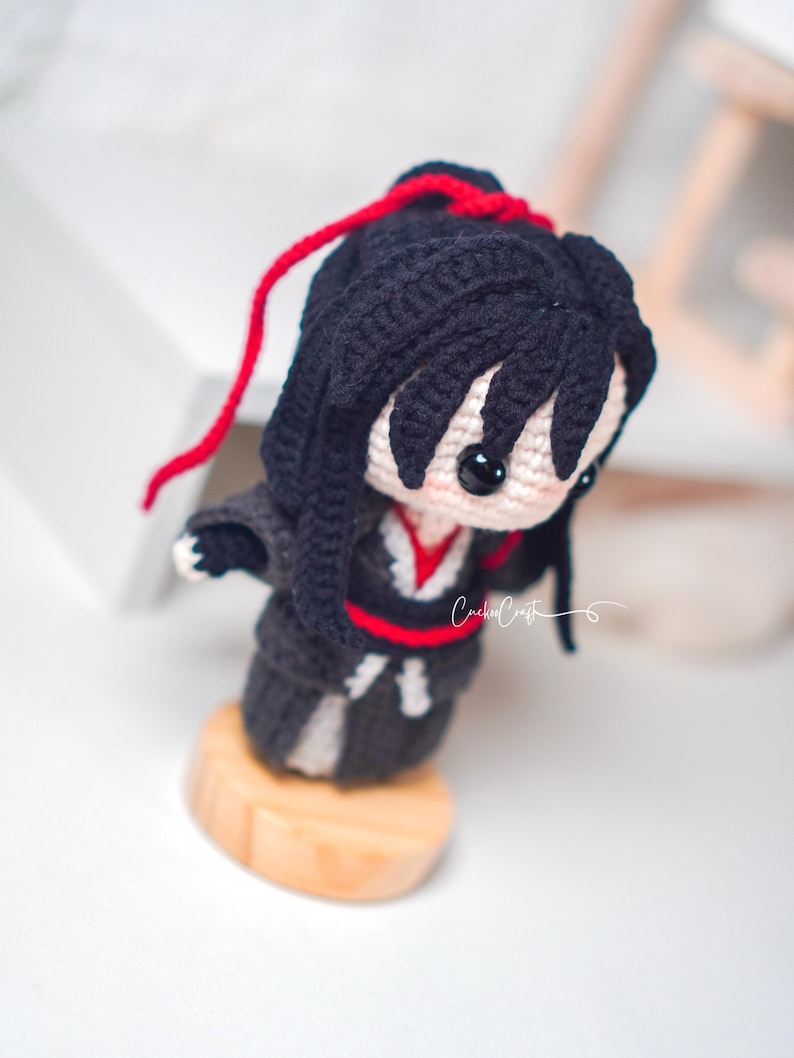 May include: Handmade crochet doll featuring black hair and a black outfit with red detailing. The doll stands on a small wooden base. It has large black eyes and a red string in its hair. The doll is a handcrafted item.