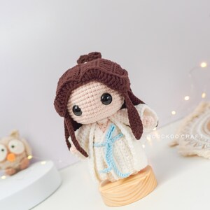 May include: A crocheted doll with brown hair, wearing a white dress with a blue sash. The doll is standing on a wooden base.
