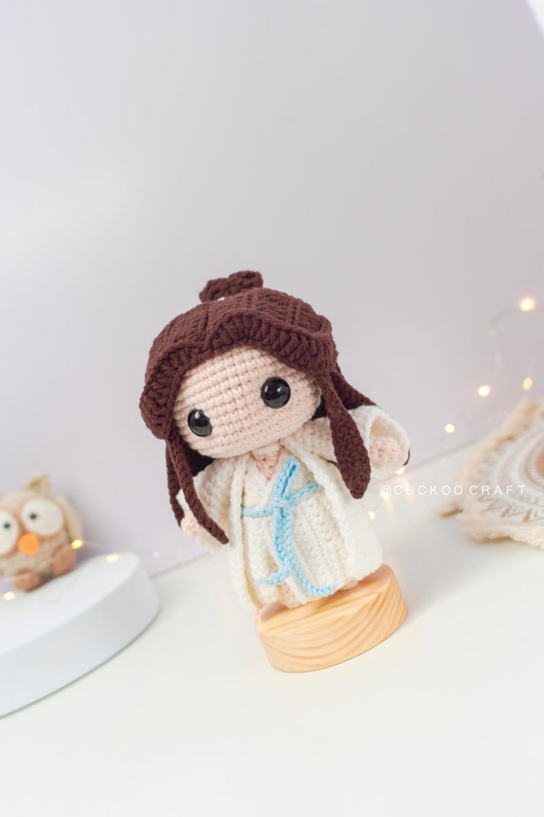 May include: A crocheted doll with brown hair, wearing a white dress with a blue sash. The doll is standing on a wooden base.