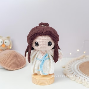 May include: A crocheted doll with brown hair, wearing a white dress with a blue sash, standing on a wooden base. The doll has large eyes and a sweet expression. The doll is surrounded by a white background.