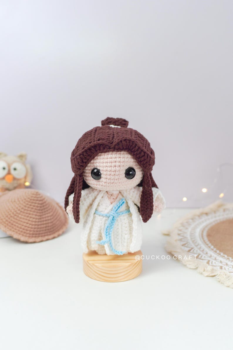 May include: A crocheted doll with brown hair, wearing a white dress with a blue sash, standing on a wooden base. The doll has large eyes and a sweet expression. The doll is surrounded by a white background.