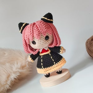 K&ouml;nnte beinhalten: Eine geh&auml;kelte Puppe mit rosa Haaren, schwarzen und gelben Kleidern und schwarzen Katzenohren. Die Puppe steht auf einem Holzsockel mit dem Text "CUKOO.CRAFT" darauf.