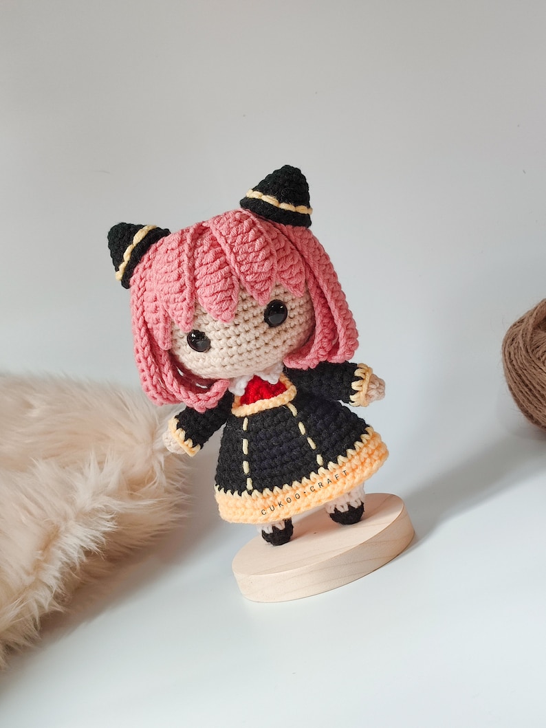 K&ouml;nnte beinhalten: Eine geh&auml;kelte Puppe mit rosa Haaren, schwarzen und gelben Kleidern und schwarzen Katzenohren. Die Puppe steht auf einem Holzsockel mit dem Text "CUKOO.CRAFT" darauf.