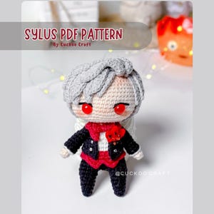 May include: A crochet doll with light gray hair, bright red eyes, and a black suit featuring a red vest and a red flower accessory. The text SYLUS PDF PATTERN By Cuckoo Craft is visible at the top, and @CUCKOO CRAFT is printed at the bottom.