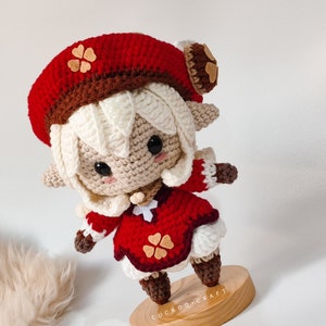 May include: Crocheted doll with white hair, a red hat with brown trim, and a red dress with brown trim. The doll is standing on a wooden base with the text "Cuckoo Craft".