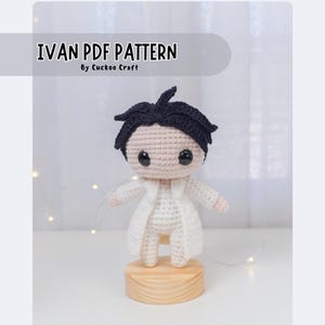 May include: Crochet amigurumi pattern for a character named Ivan. The pattern is for a white crocheted figure with black hair, wearing a white coat. The figure is standing on a wooden base. The text "IVAN PDF PATTERN by Cuckoo Craft" is displayed at the top of the image.