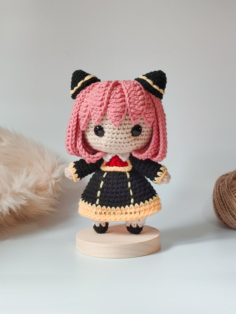 K&ouml;nnte beinhalten: Eine geh&auml;kelte Puppe mit rosa Haaren, schwarzen Ohren und einem schwarzen Kleid mit gelbem Besatz. Die Puppe steht auf einem Holzsockel. Die Puppe tr&auml;gt einen roten Kragen und hat schwarze Augen. Die Puppe ist handgefertigt und ein s&uuml;&szlig;es und fantasievolles Spielzeug.