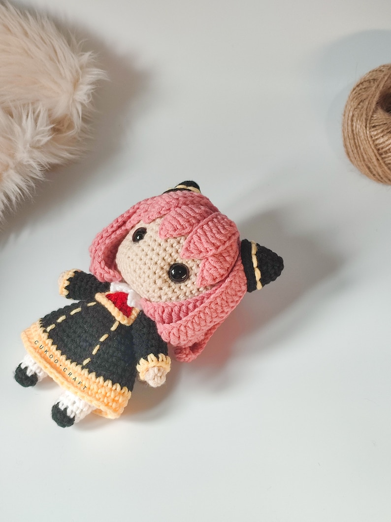 K&ouml;nnte beinhalten: Eine rosa und schwarze geh&auml;kelte Puppe mit einem schwarzen und gelben Kleid. Die Puppe hat schwarze Augen und einen kleinen, wei&szlig;en Mund. Die Puppe liegt auf einer wei&szlig;en Oberfl&auml;che. Der Text "CUKOO.CRAFT" ist auf dem Kleid der Puppe sichtbar.