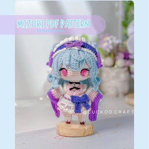 Puede incluir: Una muñeca amigurumi de ganchillo con cabello azul claro, ojos rosas y un conjunto blanco y rosa con lazos morados y una gargantilla negra. La muñeca se sostiene sobre una base de madera. El texto dice MIZUKI PDF PATTERN y @CUCKOO CRAFT.