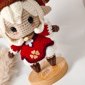 May include: A crocheted doll with white hair, a red dress, and a brown hat. The doll is standing on a wooden base with the text "Cuckoo Craft" written on it.