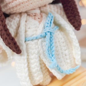 May include: Crocheted doll with brown hair wearing a white dress with a blue sash tied around the waist. The doll is standing on a wooden base. The doll's Instagram handle is @cuckoocraft.