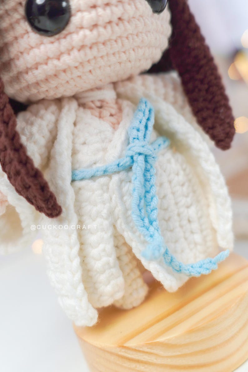May include: Crocheted doll with brown hair wearing a white dress with a blue sash tied around the waist. The doll is standing on a wooden base. The doll's Instagram handle is @cuckoocraft.