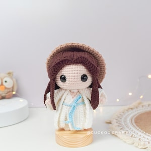 May include: A crocheted doll with brown hair, wearing a white dress and a brown hat. The doll is standing on a wooden base.