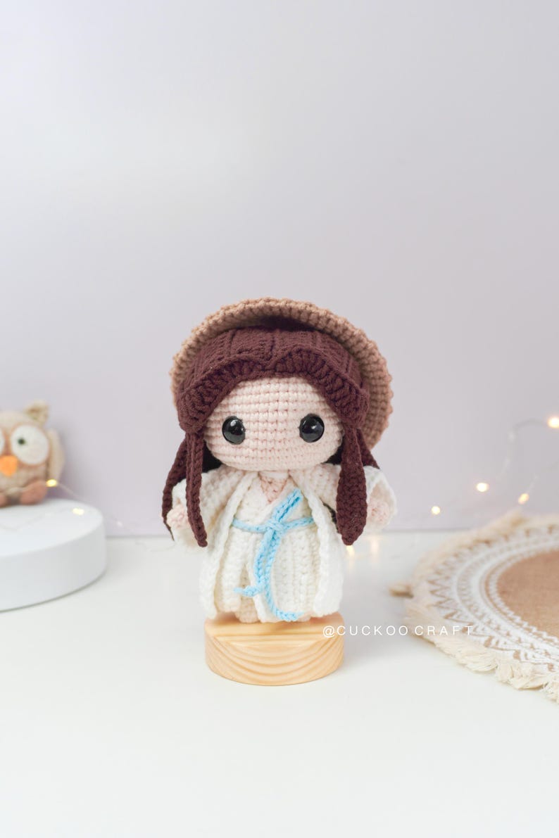 May include: A crocheted doll with brown hair, wearing a white dress and a brown hat. The doll is standing on a wooden base.