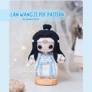 May include: A crocheted doll of Lan Wangji, a character from the novel and TV series, with black hair and a white and light blue outfit. The doll is displayed on a wooden base. The text "LAN WANGJI PDF PATTERN" is at the top.