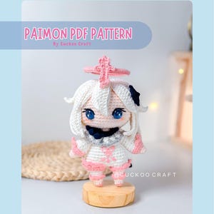 以下が含まれることがあります： 白い髪、青い目、ピンクと白の衣装が特徴の、かぎ針編みのパイモン人形。人形は小さな木製の台の上に飾られています。「Paimon PDF Pattern」の文字が人形の上に表示されています。