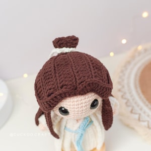 May include: A crocheted doll with brown hair, a white dress, and a blue sash. The doll is standing on a wooden base.