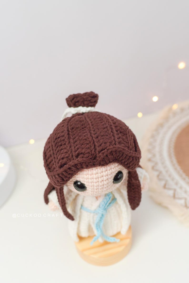 May include: A crocheted doll with brown hair, a white dress, and a blue sash. The doll is standing on a wooden base.