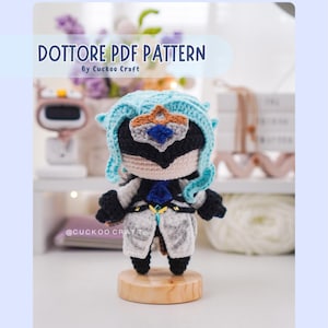 Puede incluir: Muñeca de ganchillo hecha a mano de un personaje con máscara negra, pelo azul y atuendo blanco y gris. La muñeca está sobre una base de madera. El texto "Dottore PDF Pattern" aparece en la parte superior de la imagen.
