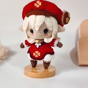 May include: A crocheted doll with white hair, a red hat with brown trim and a red dress with white trim. The doll is standing on a wooden base with the text "Cuckoo Craft".