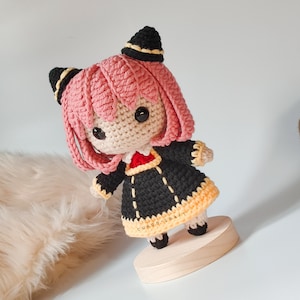 K&ouml;nnte beinhalten: Eine geh&auml;kelte Amigurumi-Puppe mit rosa Haaren, schwarzer und gelber Kleidung und schwarzen Augen. Die Puppe steht auf einem Holzsockel. Der Text "CUKOO.CRAFT" ist auf dem Sockel sichtbar.