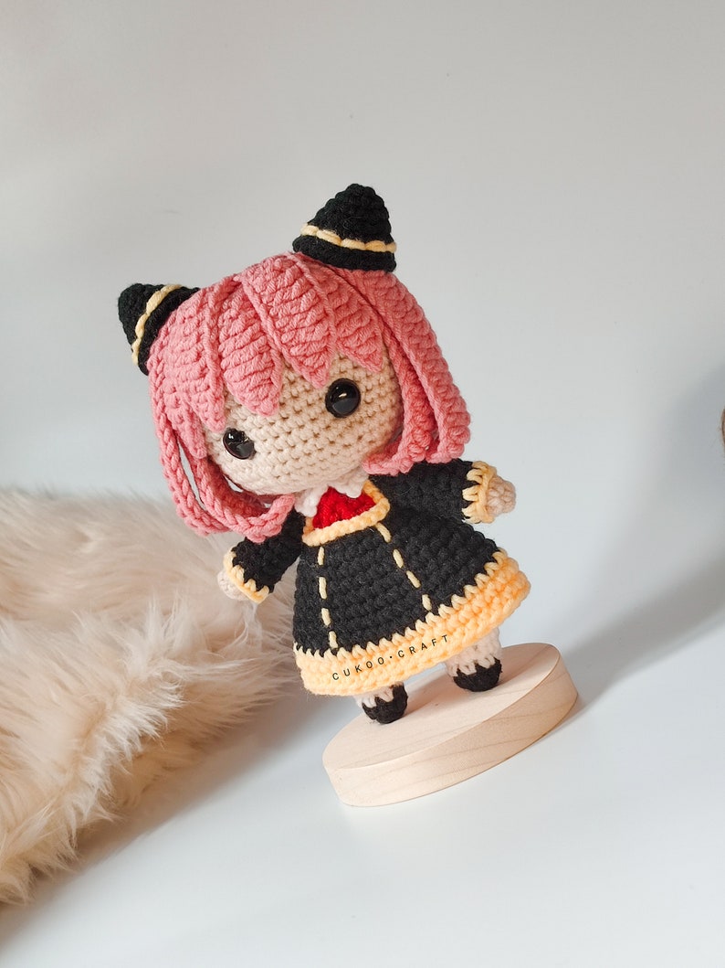 K&ouml;nnte beinhalten: Eine geh&auml;kelte Amigurumi-Puppe mit rosa Haaren, schwarzer und gelber Kleidung und schwarzen Augen. Die Puppe steht auf einem Holzsockel. Der Text "CUKOO.CRAFT" ist auf dem Sockel sichtbar.