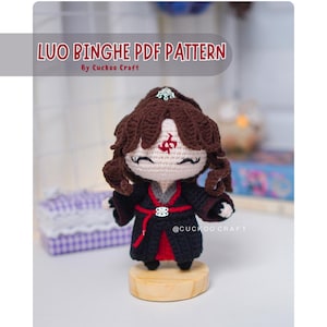 Puede incluir: Muñeca de ganchillo de un personaje con cabello rizado castaño, vestida con un atuendo negro y rojo. La muñeca está sobre una base de madera. El texto "LUO BINGHE PDF PATTERN" está en la parte superior, con "By Cuckoo Craft" debajo.