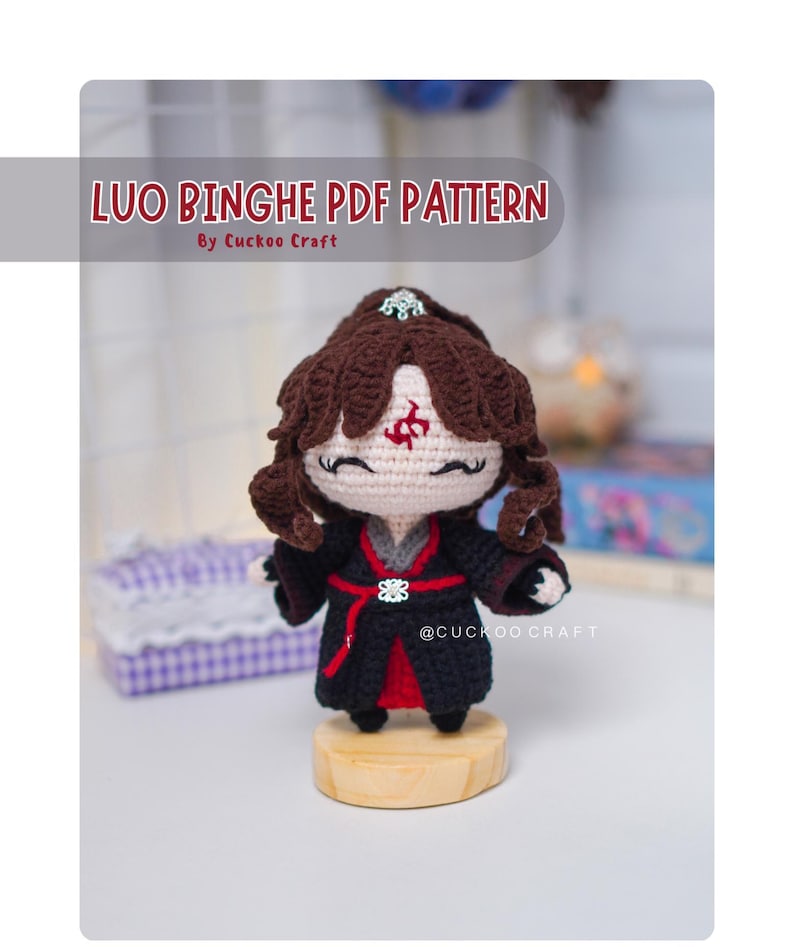 Op de afbeelding: Gehaakte pop van een personage met bruin krullend haar, gekleed in een zwart en rode outfit. De pop staat op een houten voet. De tekst "LUO BINGHE PDF PATTERN" staat bovenaan, met "By Cuckoo Craft" eronder.