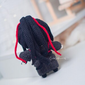 May include: Handmade crochet doll with black hair and red strands. The doll is dressed in a dark grey outfit and has small black shoes. The doll has a unique design.