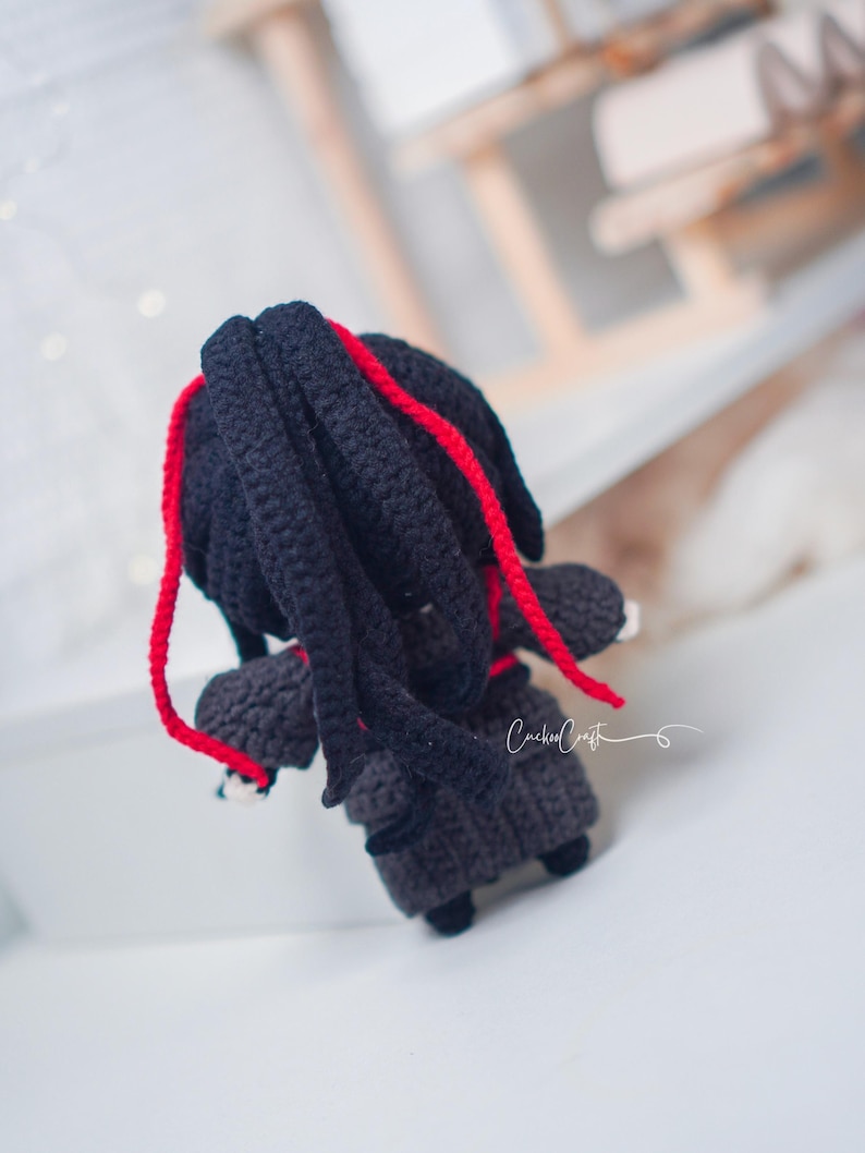 May include: Handmade crochet doll with black hair and red strands. The doll is dressed in a dark grey outfit and has small black shoes. The doll has a unique design.