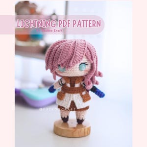 以下が含まれることがあります： ピンクの髪、青い目、茶色と白の衣装のキャラクターの手作りの編み人形。人形は木の台座の上に立っています。画像には「LIGHTNING PDF PATTERN」というテキストが表示されています。