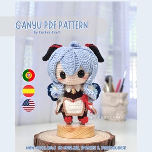 Puede incluir: Muñeca amigurumi de ganchillo de Ganyu del videojuego Genshin Impact. La muñeca lleva un vestido azul y blanco con una cinta roja. La muñeca está de pie sobre una base de madera. La imagen incluye el texto "GANYU PDF PATTERN" y "NOW AVAILABLE IN ENGLISH, SPANISH & PORTUGUESE".
