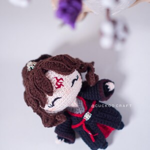 Op de afbeelding: Een handgemaakte gehaakte pop met bruin haar en een zwart-rode outfit. De pop heeft een wit gezicht met gesloten ogen en een rood symbool op het voorhoofd. De tekst "@CUCKOO CRAFT" is zichtbaar op de kleding.