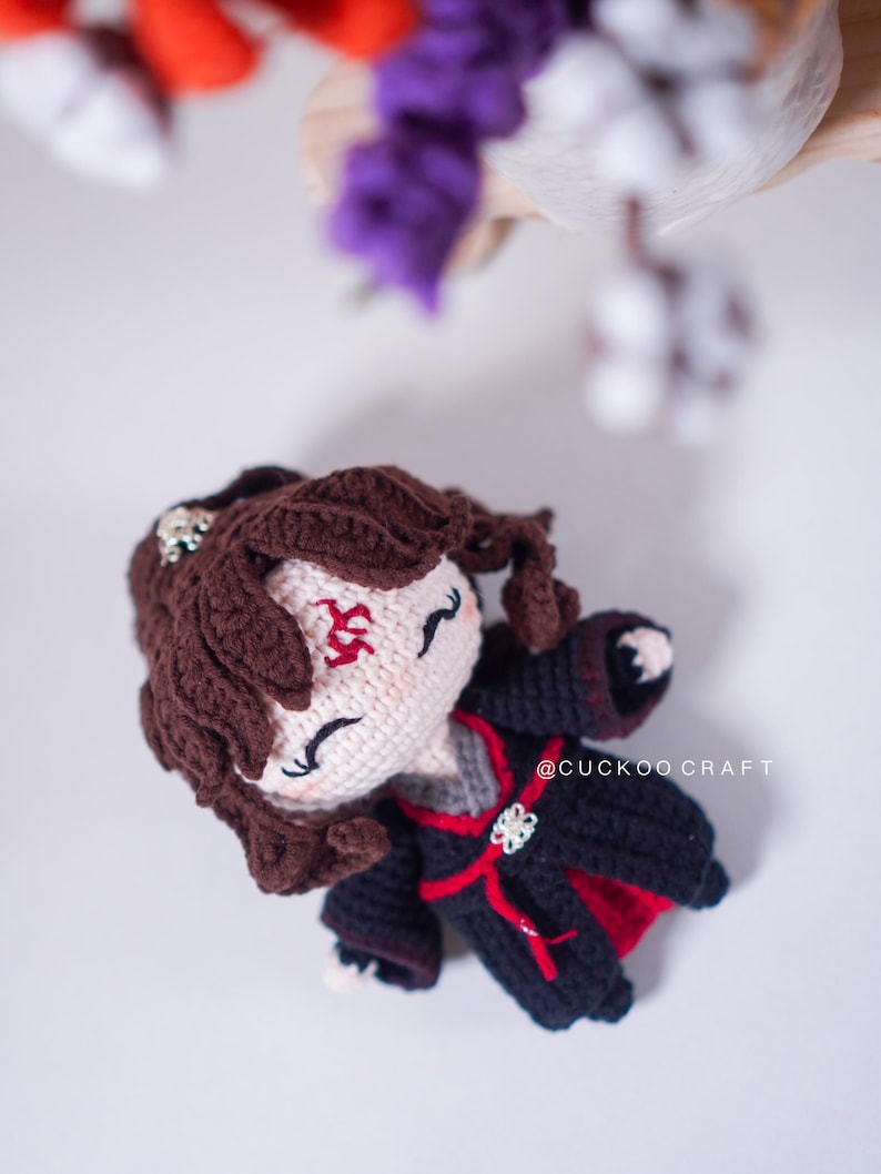 Op de afbeelding: Een handgemaakte gehaakte pop met bruin haar en een zwart-rode outfit. De pop heeft een wit gezicht met gesloten ogen en een rood symbool op het voorhoofd. De tekst "@CUCKOO CRAFT" is zichtbaar op de kleding.