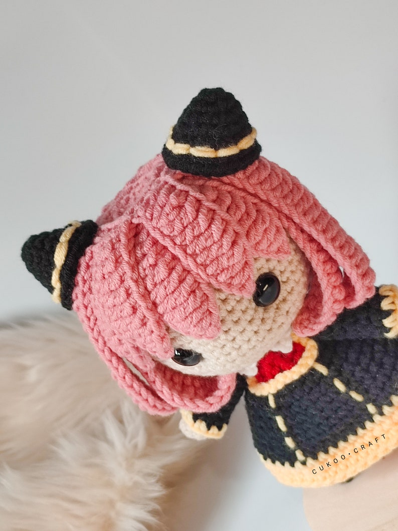 K&ouml;nnte beinhalten: Eine geh&auml;kelte Puppe mit rosa Haaren, schwarzen H&ouml;rnern und einem schwarzen und gelben Hut. Die Puppe tr&auml;gt ein schwarzes und gelbes Kleid. Die Puppe ist handgefertigt und hat einen s&uuml;&szlig;en Ausdruck.