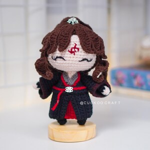 Op de afbeelding: Gehaakte pop met bruin haar en een zwart en rode outfit. De pop heeft een rood symbool op het voorhoofd en staat op een kleine houten basis. De tekst "@CUCKOO CRAFT" is zichtbaar.