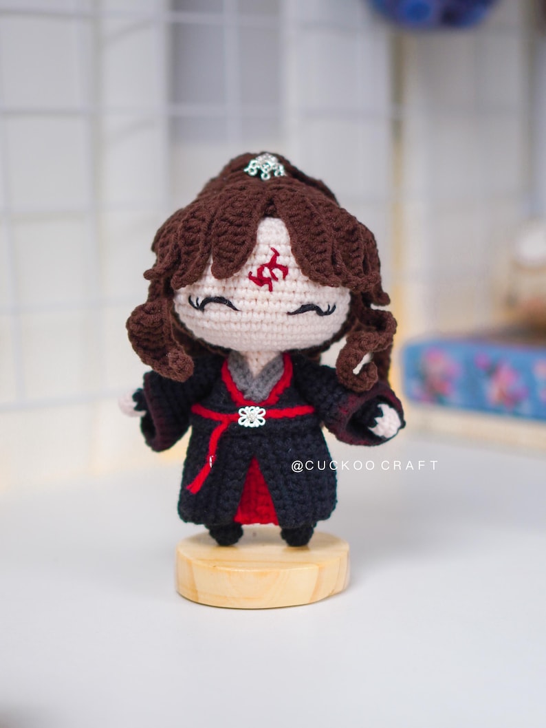 Op de afbeelding: Gehaakte pop met bruin haar en een zwart en rode outfit. De pop heeft een rood symbool op het voorhoofd en staat op een kleine houten basis. De tekst "@CUCKOO CRAFT" is zichtbaar.