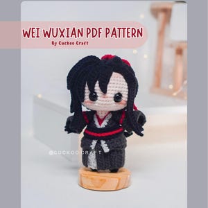 May include: A crocheted doll of a character with black hair and a red hair accessory, wearing a black and gray outfit with red accents. The doll is on a wooden base. Text on the image reads "WEI WUXIAN PDF PATTERN" and "By Cuckoo Craft".