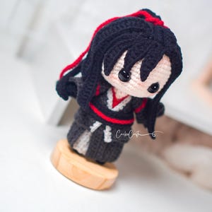 May include: A crocheted doll with black hair and red accents, dressed in a black and grey outfit with red trim. The doll is mounted on a small wooden base. The doll has black button eyes and is a unique handcrafted item.