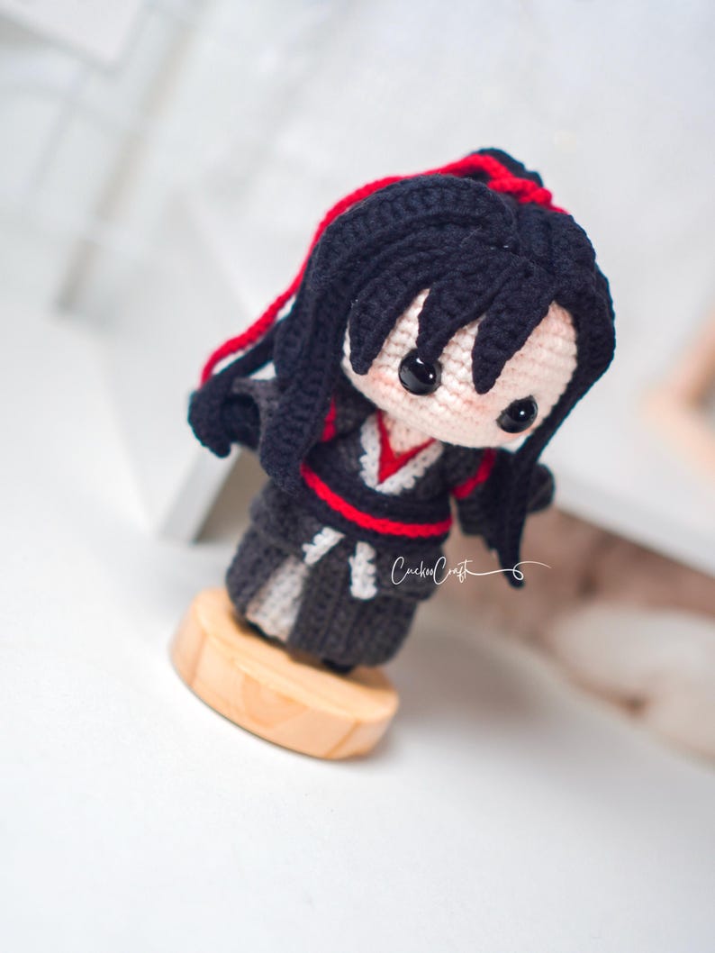 May include: A crocheted doll with black hair and red accents, dressed in a black and grey outfit with red trim. The doll is mounted on a small wooden base. The doll has black button eyes and is a unique handcrafted item.