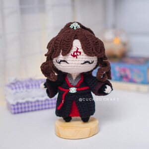 Op de afbeelding: Gehaakte pop met bruin haar en een zwarte outfit met rode accenten. De pop heeft een decoratieve hoofdtooi en een rood symbool op het voorhoofd. Hij staat op een kleine houten basis. Handgemaakt ambacht.