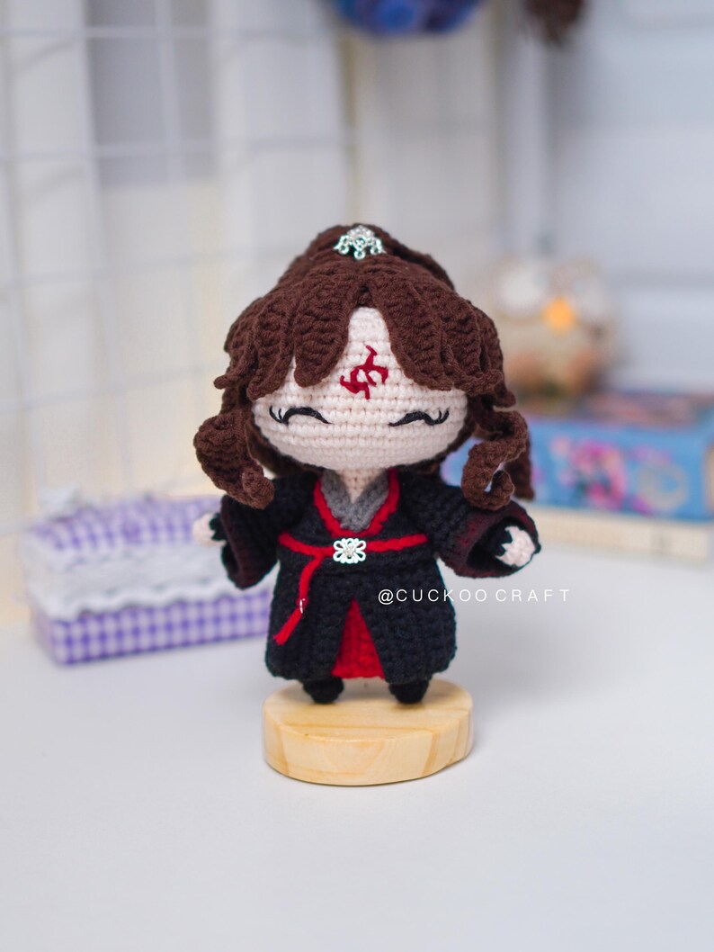 Op de afbeelding: Gehaakte pop met bruin haar en een zwarte outfit met rode accenten. De pop heeft een decoratieve hoofdtooi en een rood symbool op het voorhoofd. Hij staat op een kleine houten basis. Handgemaakt ambacht.