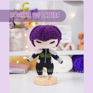 May include: A handmade amigurumi doll with a purple hair, black and gray outfit, and a wooden base. The doll has a neutral expression and is displayed against a soft background. The text "HOSHINA PDF PATTERN" is visible.