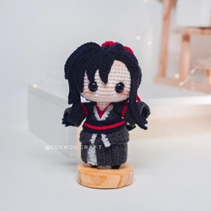 May include: A crocheted doll with black hair and a black and red outfit, standing on a small wooden base. The doll has a light beige face with black eyes. The text "@CUCKOOCRAFT" is visible on the base. The doll is a handmade item.